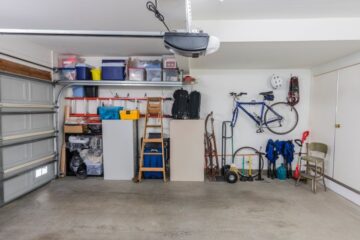 A clean garage with white walls and fluorescent lighting shows a clean interior with clear, organized bins on the shelf.