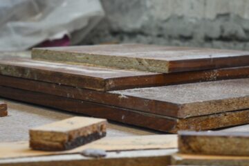 Dark-stained wooden planks and untreated scraps of different sizes rest in stacks on the ground near a concrete wall.