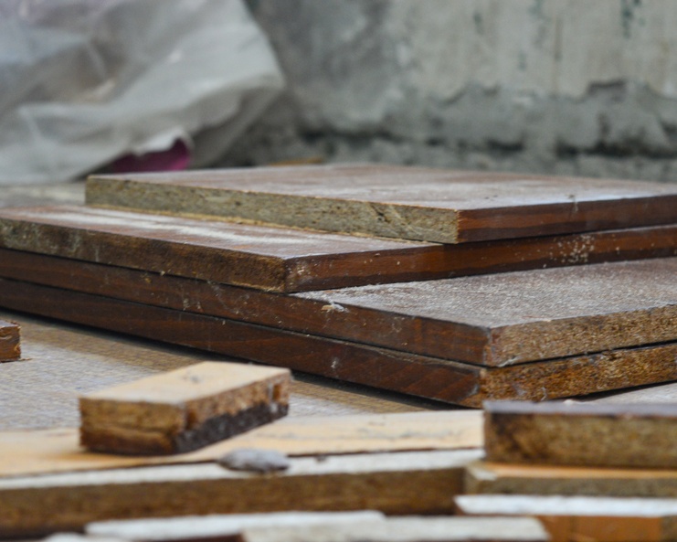 Dark-stained wooden planks and untreated scraps of different sizes rest in stacks on the ground near a concrete wall.