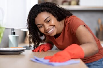 A smiling woman wearing an orange shirt and rubber gloves wipes a wooden kitchen counter with a blue cloth.
