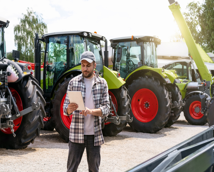 A man wearing a plaid shirt and a baseball cap holds a tablet while standing in front of a row of green tractors.
