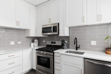 A small kitchen with white cabinets with gold hardware, gray backsplash tiles, and stainless steel appliances.