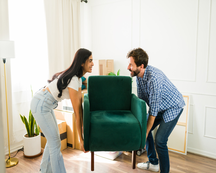 A couple smiling at each other as they carry a velvet green reading chair into a corner in their home.