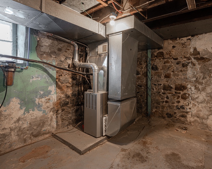 A modern HVAC system with metal ductwork sits in the corner of a residential basement near an exposed stone wall.