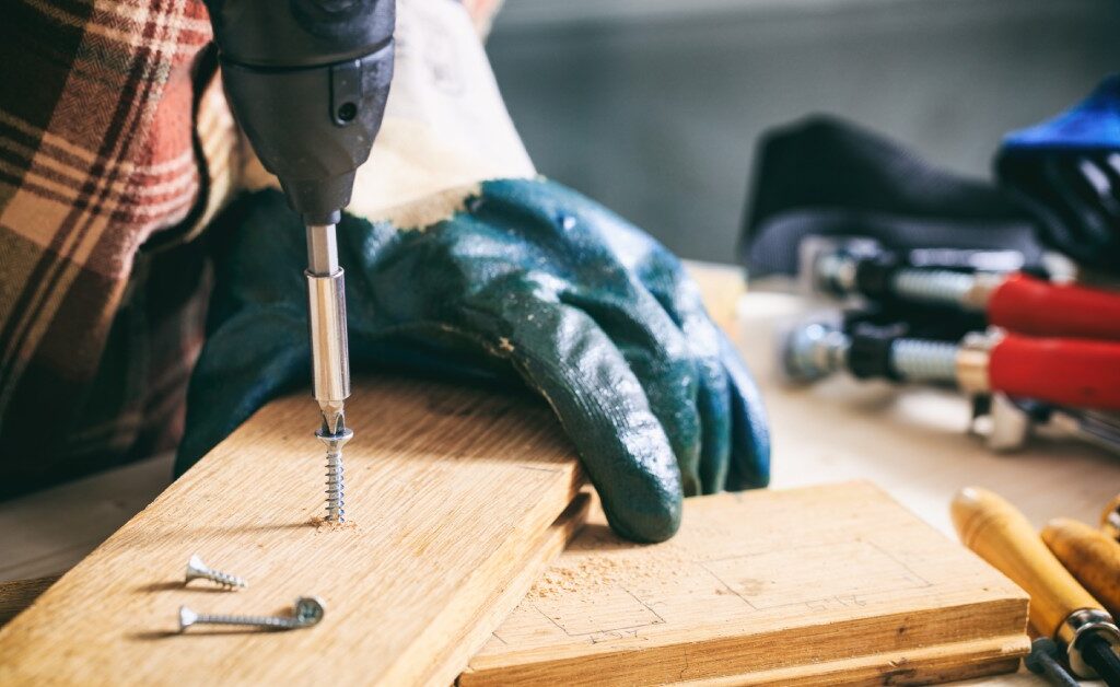 A person uses a power drill to drive a screw into a narrow piece of wood. Tools surround the project on a workshop table.