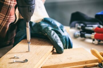 A person uses a power drill to drive a screw into a narrow piece of wood. Tools surround the project on a workshop table.