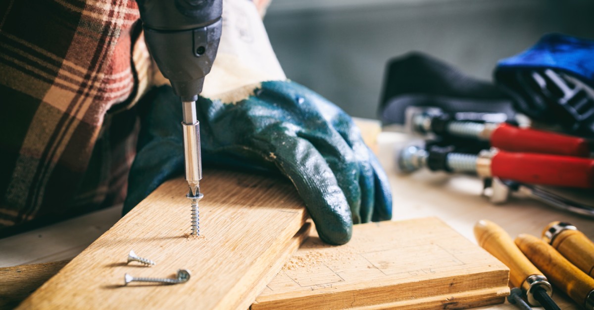 A person uses a power drill to drive a screw into a narrow piece of wood. Tools surround the project on a workshop table.
