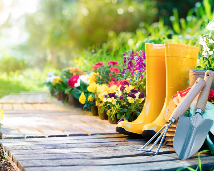 A wooden walkway on the ground surrounded by flowers next to a pair of yellow boots, and various gardening tools.