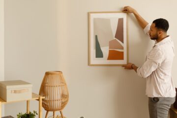 A young Black man in a dress shirt and pants carefully hangs a framed piece of art on a clean, white wall.