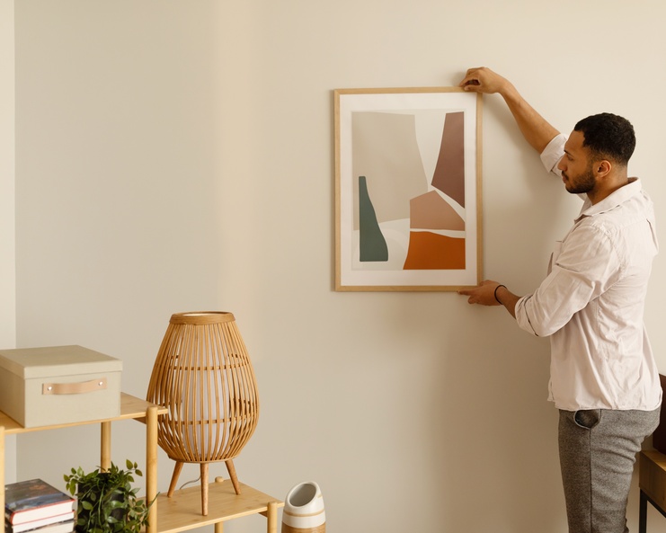 A young Black man in a dress shirt and pants carefully hangs a framed piece of art on a clean, white wall.