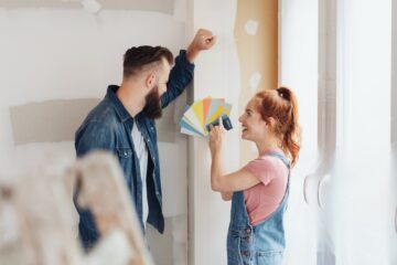 A woman and man stand in front of a half-painted wall. The woman grins as she holds up paint swatches.