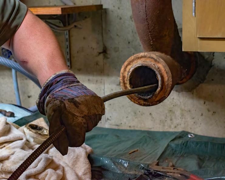 A person wearing a thick brown glove as they guide a drain snake into a rusted pipe with a green tarp underneath.