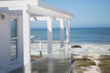 A waterfront property built next to a beach. The home has a balcony with glass railings and a white roofing structure.
