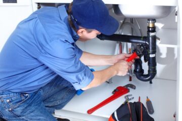 A person wearing a blue shirt, jeans, and cap uses a crescent wrench to tighten the pipes underneath a sink.