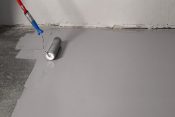 A paint roller spreads gray epoxy coating evenly across a concrete floor next to a white wall without a baseboard.