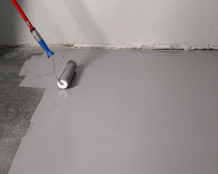 A paint roller spreads gray epoxy coating evenly across a concrete floor next to a white wall without a baseboard.