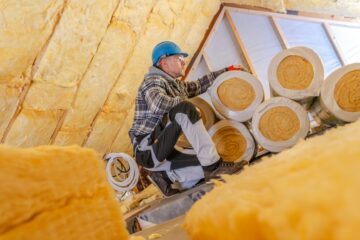 A worker kneeling in an attic installing rolls of insulation. He is wearing a jacket and a hard hat.