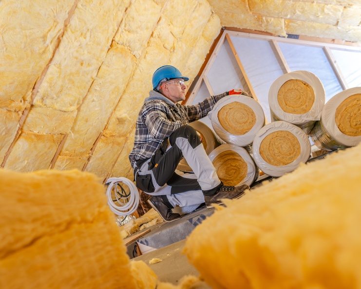 A worker kneeling in an attic installing rolls of insulation. He is wearing a jacket and a hard hat.