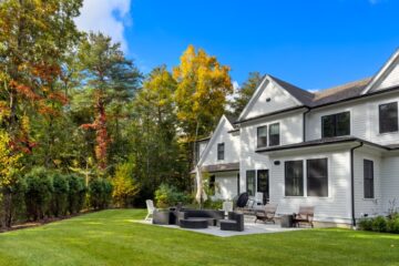 A white house standing behind a patio with black wicker furniture and a yard lined with green grass, shrubbery, and trees.