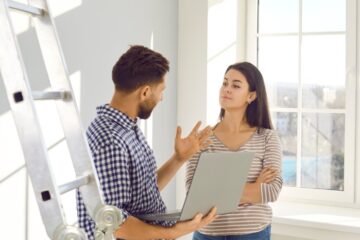 A man wearing a leather toolbelt talking to a woman inside a house. The man is holding a laptop in his hands.