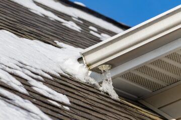 A brown asphalt shingle roof with patches of snow surrounding a white gutter with small icicles hanging from the end.