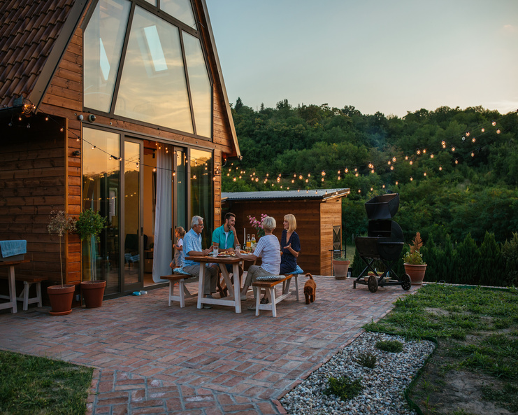 Four people sitting on two benches around a wooden table in a backyard patio with a grill and string lights.