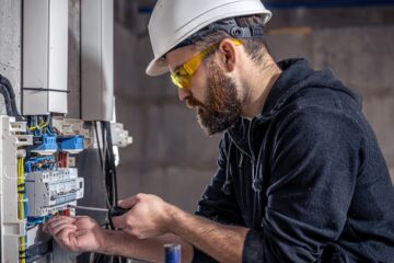 An electrician wearing a hard hat and safety goggles uses a tool to work on an electrical panel with various wires.
