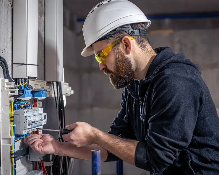 An electrician wearing a hard hat and safety goggles uses a tool to work on an electrical panel with various wires.