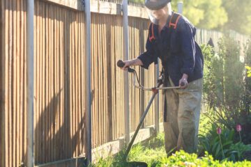 A woman wearing a face guard while using a brush cutter. She is cutting the long grass next to her fence.