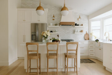 A kitchen with white cabinets, subway tile backsplash, wooden bar stools, and brass pendant lights over a white island.