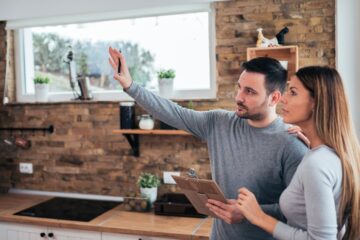 A young couple looking at their home, the man holding a clipboard and gesturing to a part of the home off screen.