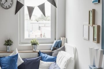 A small window in a bedroom with a shelf under it and a bed against the wall. There's a hanging banner over the window.