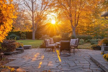 The sun peeks in between the autumn trees and illuminates the gray outdoor furniture sitting on top of a stone patio.