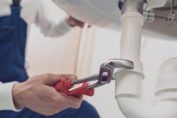 A plumber wearing a white shirt and blue overalls crouches down and uses a pipe wrench to fix the sink.
