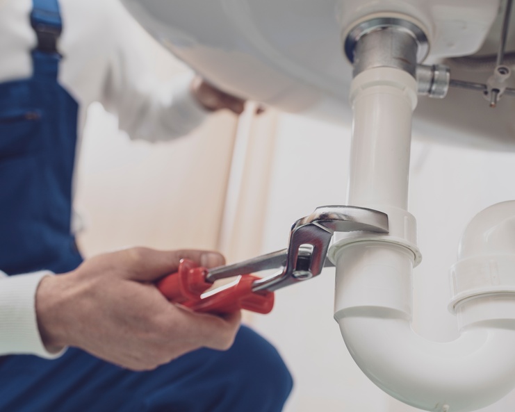 A plumber wearing a white shirt and blue overalls crouches down and uses a pipe wrench to fix the sink.