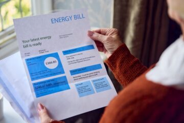 A closeup of an older woman wearing an orange sweater holding an energy bill. Behind it is a white cup of coffee.