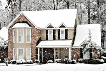 Snow blankets a two-story brick house as flakes fall steadily, coating the roof, trees, and front yard.