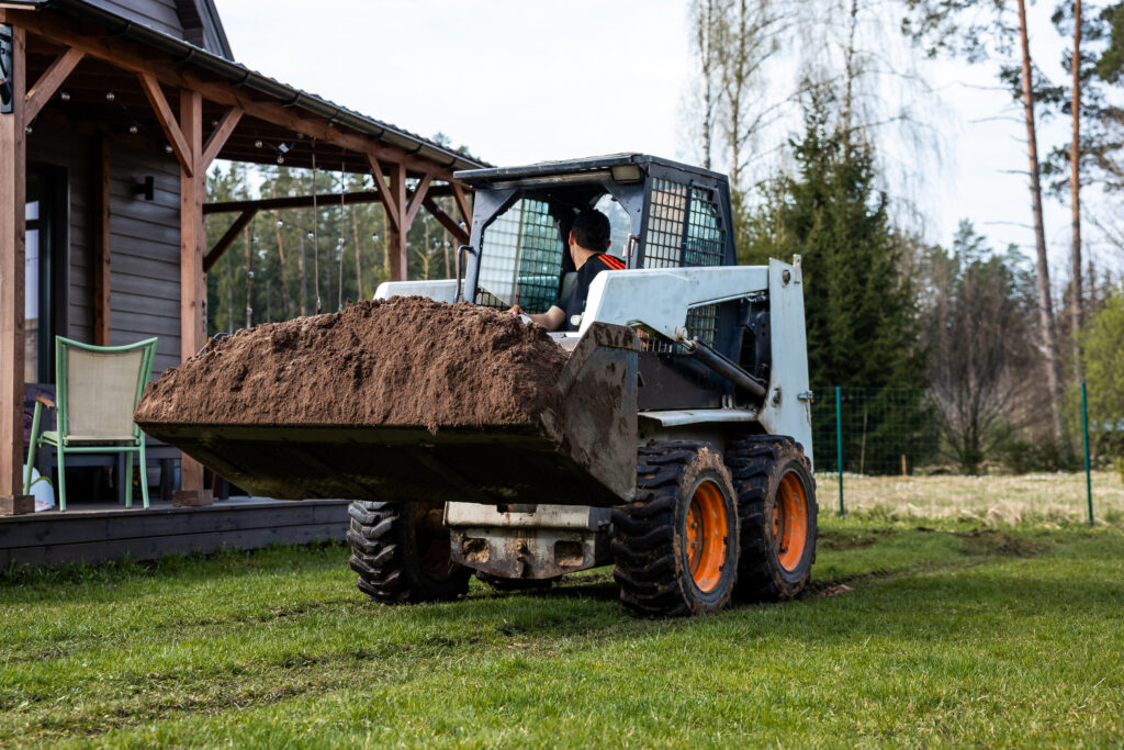 An operator looking backward while driving a skid steer next to a house. The skid steer has a bucket attachment full of dirt.