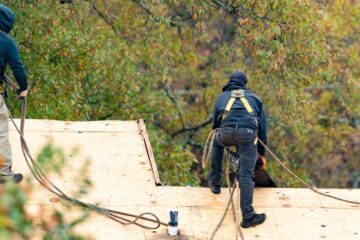Two roofers in black hoodies and yellow harnesses stand on a home's exposed roof deck under a tree.
