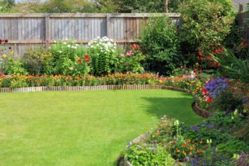 Colorful shrubs and bushes grow in front of a wooden panel fence, adding layered greenery and bright foliage contrast.