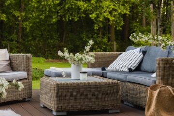 A backyard set of wicker furniture with blue and white pillows on each seat. There are white flowers around the chairs.