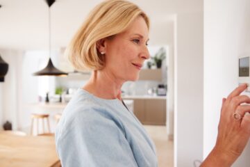 A woman with short blonde hair, earrings, and a blue top touches a wall-mounted thermostat in a home.