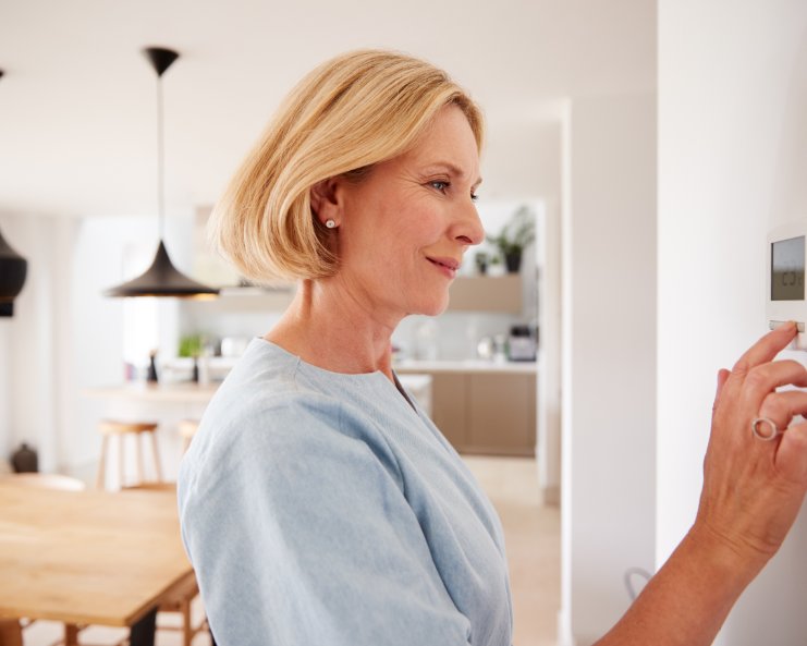 A woman with short blonde hair, earrings, and a blue top touches a wall-mounted thermostat in a home.
