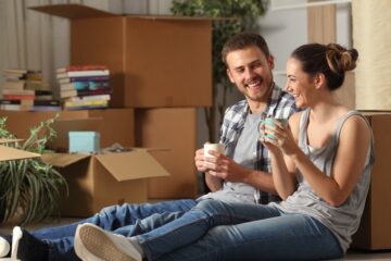 A couple sits on the floor enjoying tea in mugs as they smile at each other with moving boxes around them.