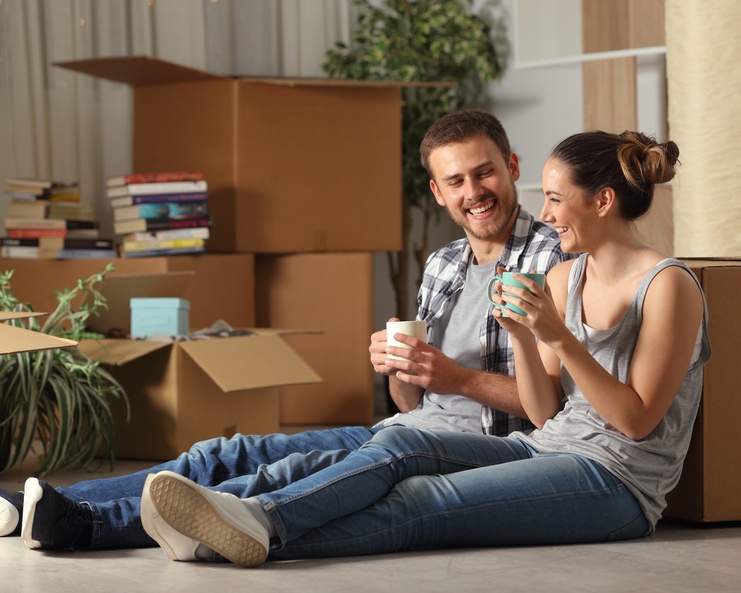 A couple sits on the floor enjoying tea in mugs as they smile at each other with moving boxes around them.