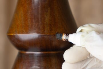 A person wearing a white glove uses a wood stain touch-up marker on the surface of a wood spindle for restoration work.