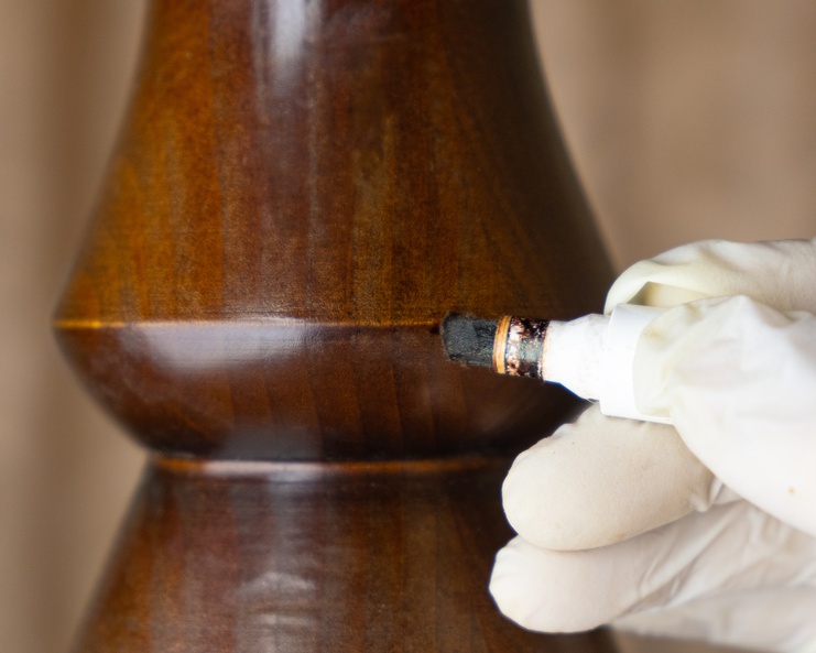 A person wearing a white glove uses a wood stain touch-up marker on the surface of a wood spindle for restoration work.