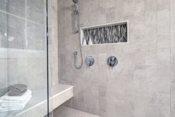 A large walk-in shower with a bench and a small niche for bottles. The tile is grey, and the shower has a removable head.