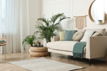 A living room has a beige sofa topped with patterned pillows and a throw blanket. A macrame wall hanging is above it.