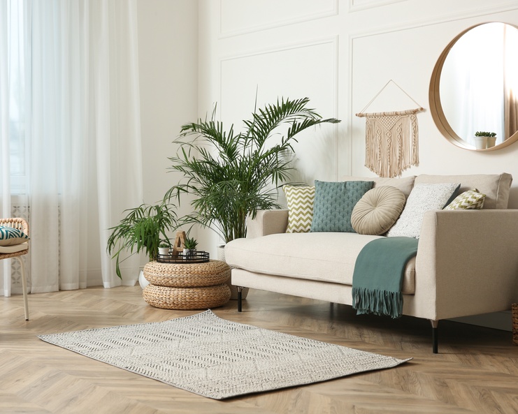 A living room has a beige sofa topped with patterned pillows and a throw blanket. A macrame wall hanging is above it.
